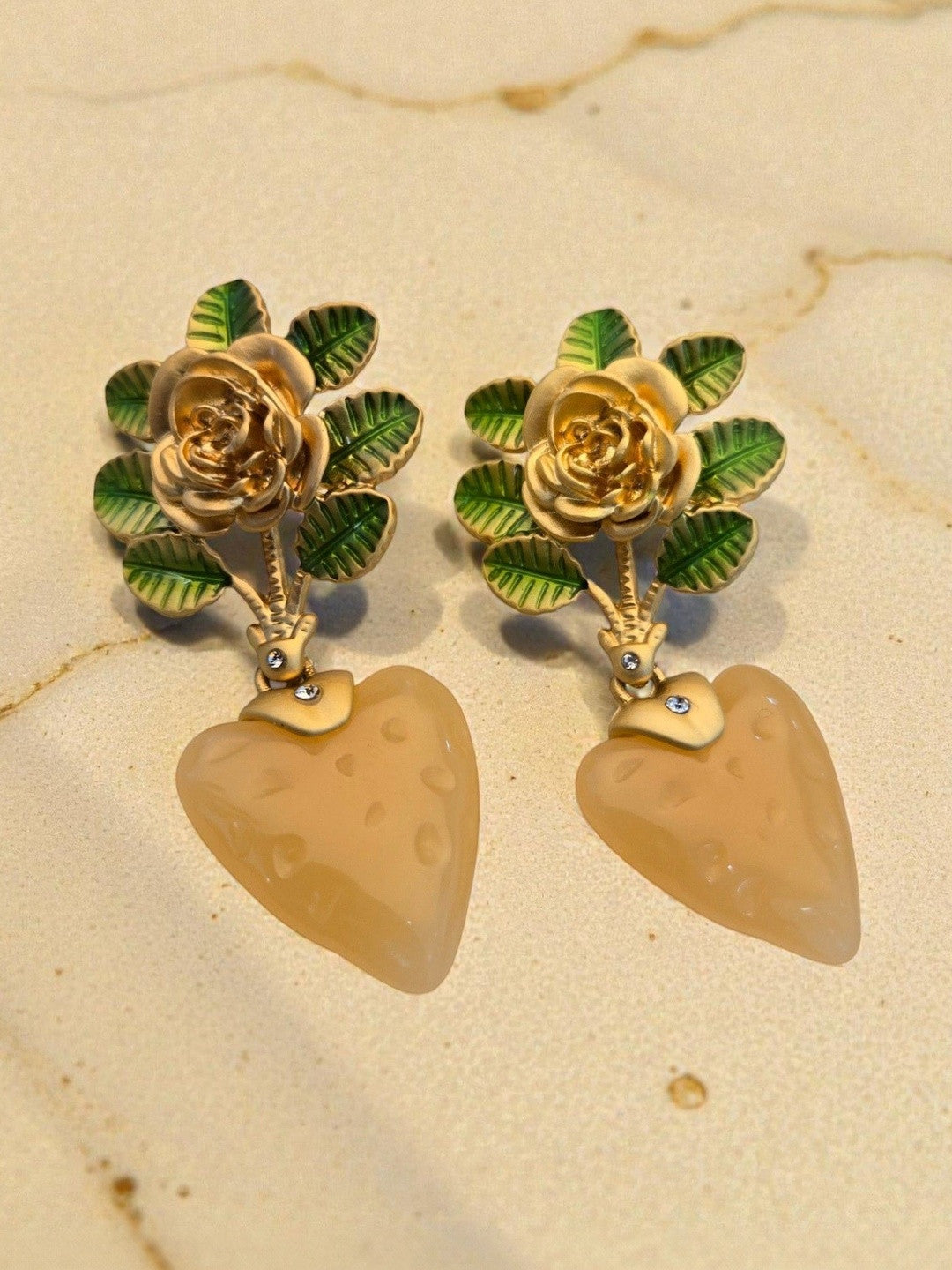 Close-up of gold rose and enamel leaf earrings with dimpled peach resin heart drops resting on a beige marble surface