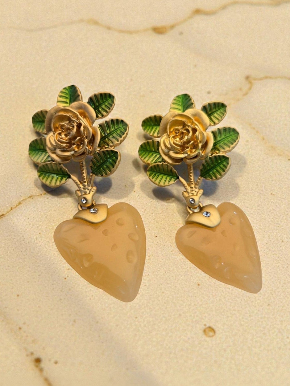 Close-up of gold rose and enamel leaf earrings with dimpled peach resin heart drops resting on a beige marble surface