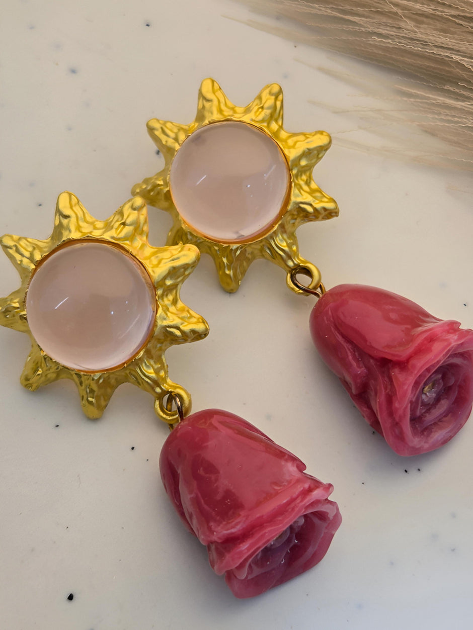Two gold-plated sun stud earrings with pink cabochons and deep pink resin rose drops, laid flat on a white surface
