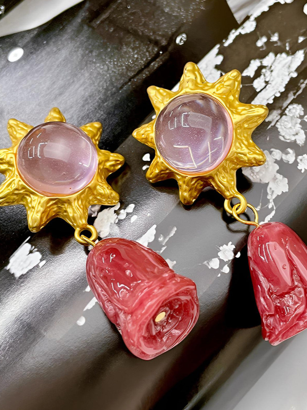 Close-up of two gold sun earrings with translucent pink cabochons and textured resin rose pendants on a dark surface