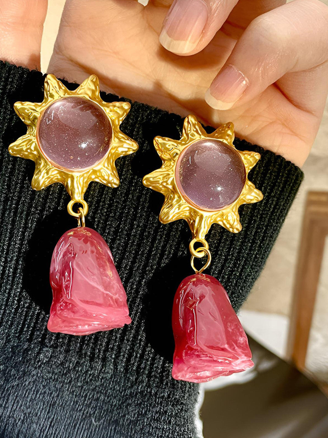 Gold-plated sun stud earrings with pink cabochons and translucent pink resin rose drops held against a black ribbed sweater