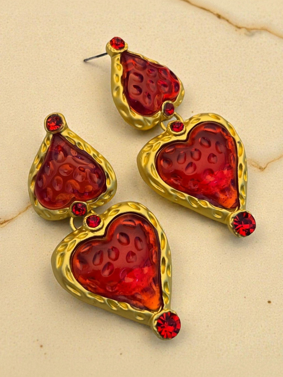 Pair of gold-tone double heart drop earrings with red resin inserts and crystal accents on a beige marble surface