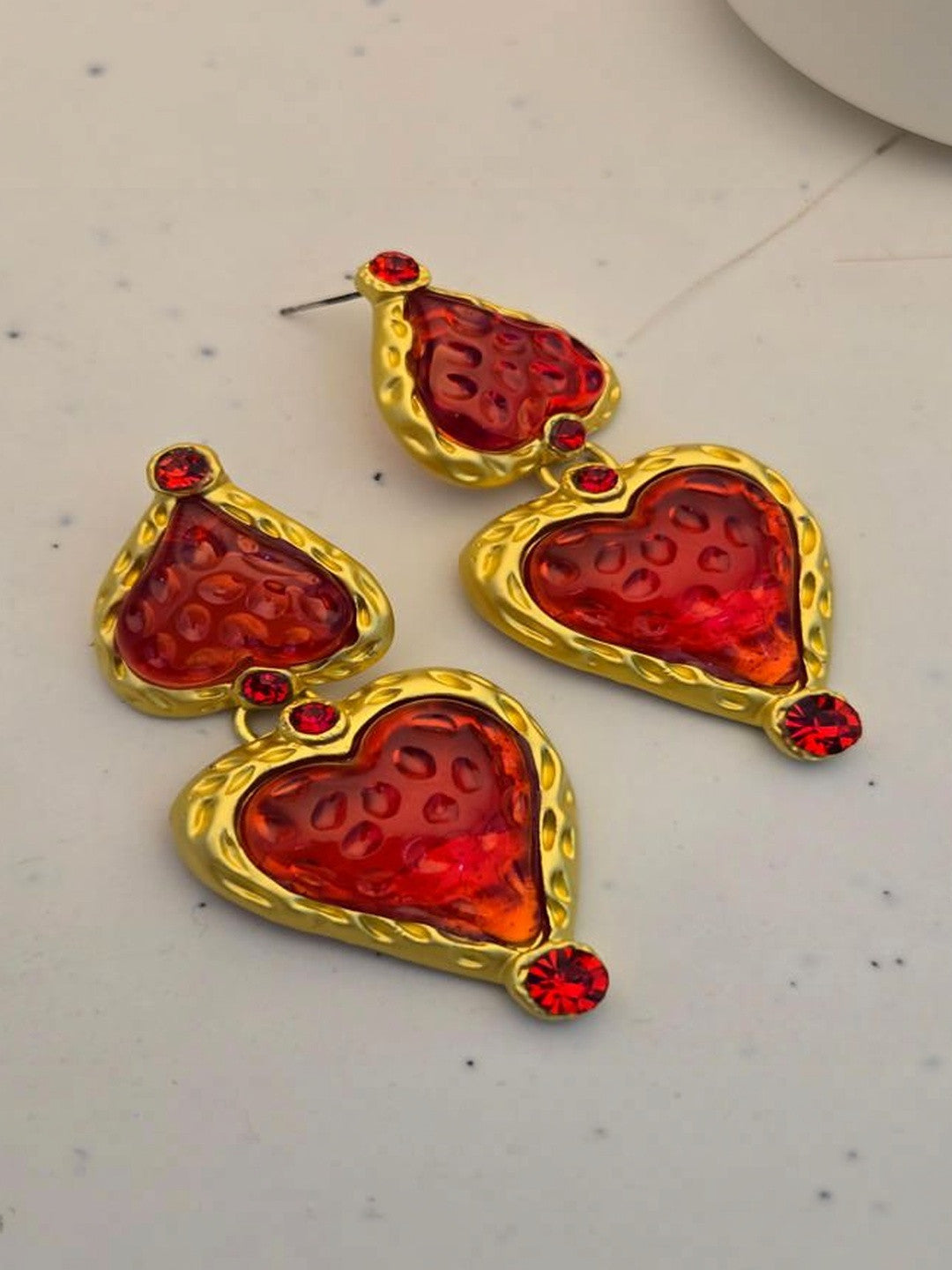 Both gold-tone red resin heart drop earrings laid flat on a white marble surface showing textured frame detail
