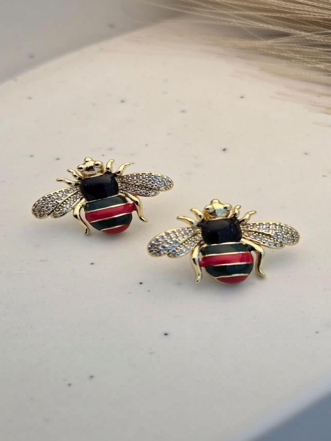 Pair of gold-plated bee stud earrings with red and green enamel abdomen and crystal wings on white surface