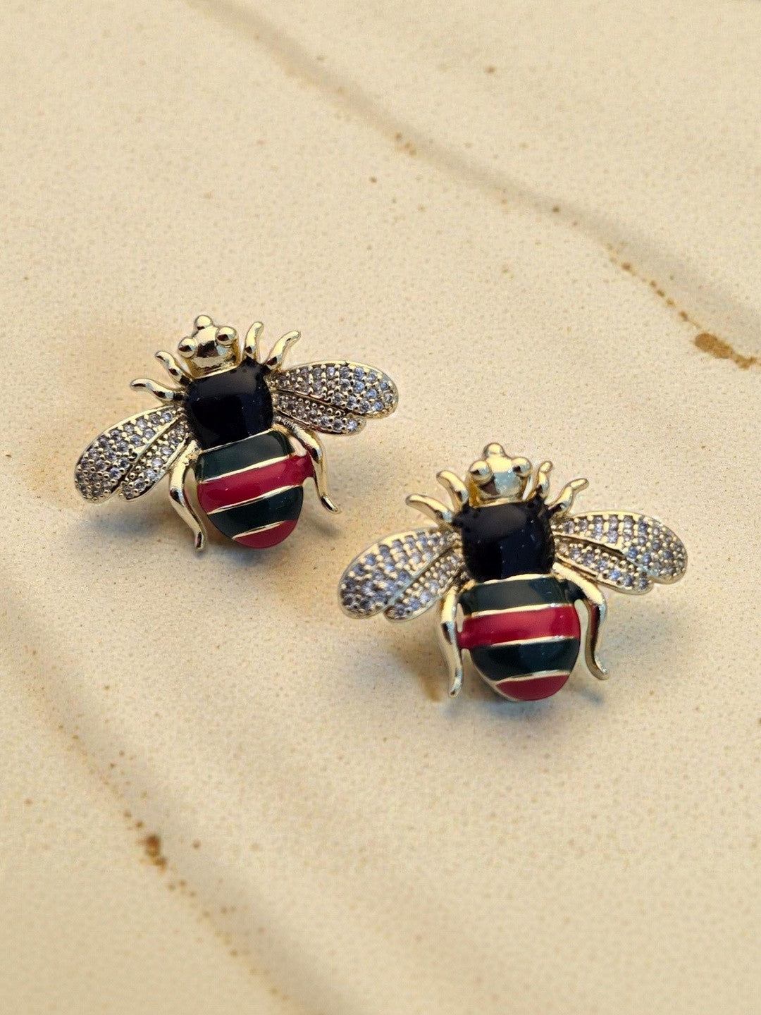 Close-up of gold-plated bee stud earring pair on sandy beige surface showing enamel and crystal detail