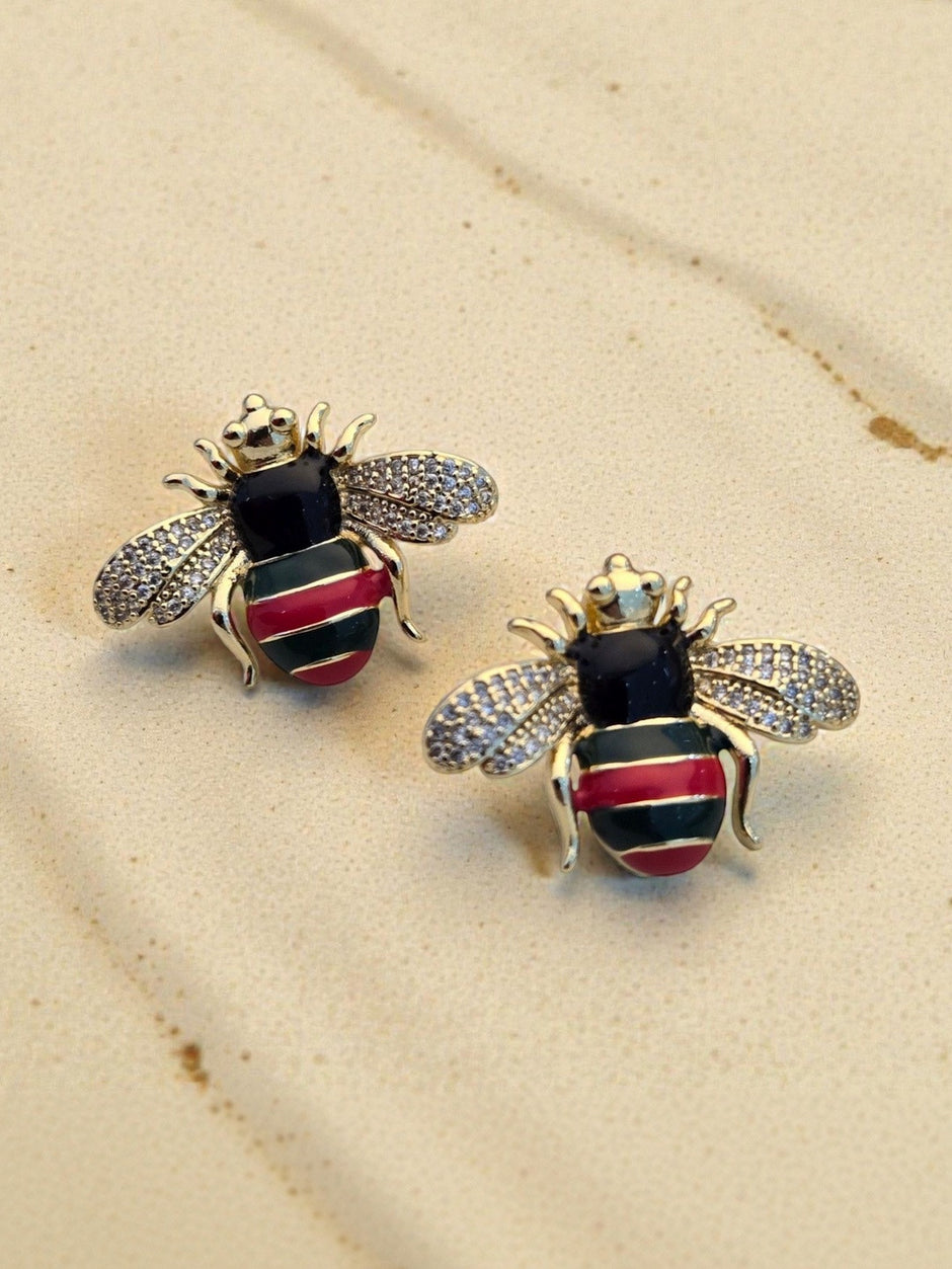 Close-up of gold-plated bee stud earring pair on sandy beige surface showing enamel and crystal detail