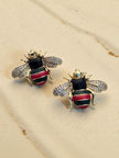 Close-up of gold-plated bee stud earring pair on sandy beige surface showing enamel and crystal detail