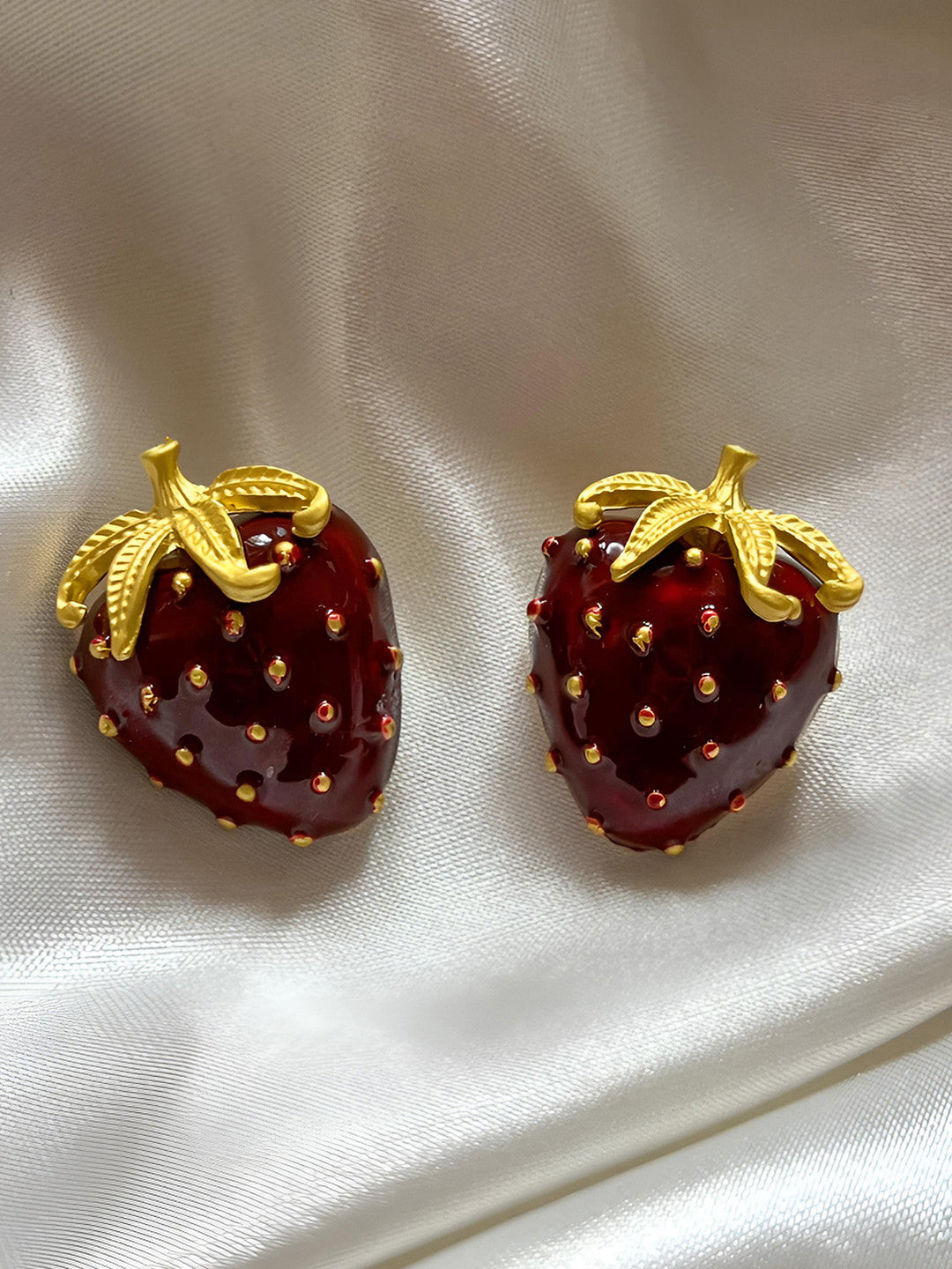 Pair of red enamel strawberry stud earrings with gold-plated leaves on white satin fabric