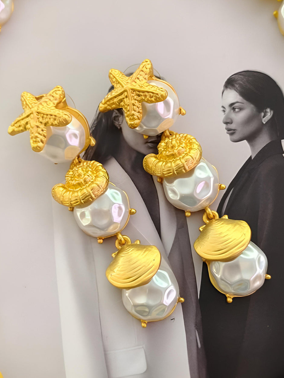 Two gold-plated drop earrings laid flat on a fashion magazine, showing starfish, nautilus and clam shell tiers with baroque faux pearls