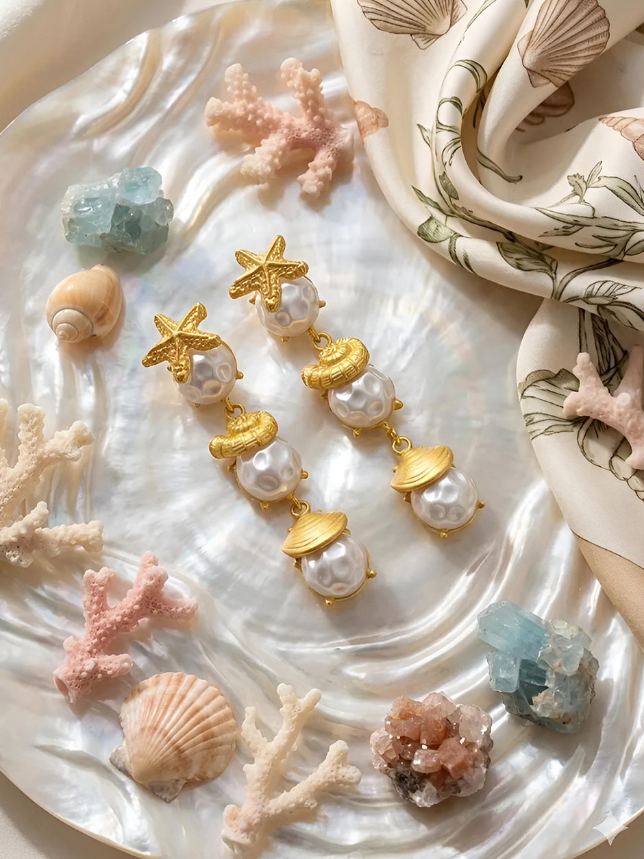 Pair of gold sea shell pearl drop earrings displayed on a mother-of-pearl shell surrounded by coral and aquamarine crystals