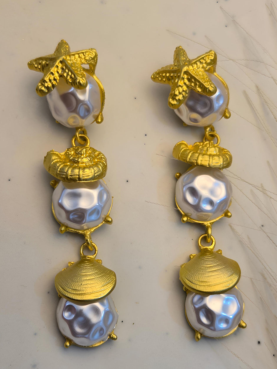 Close-up flat lay of both gold-plated sea shell drop earrings on white marble showing starfish, nautilus and clam shell detail