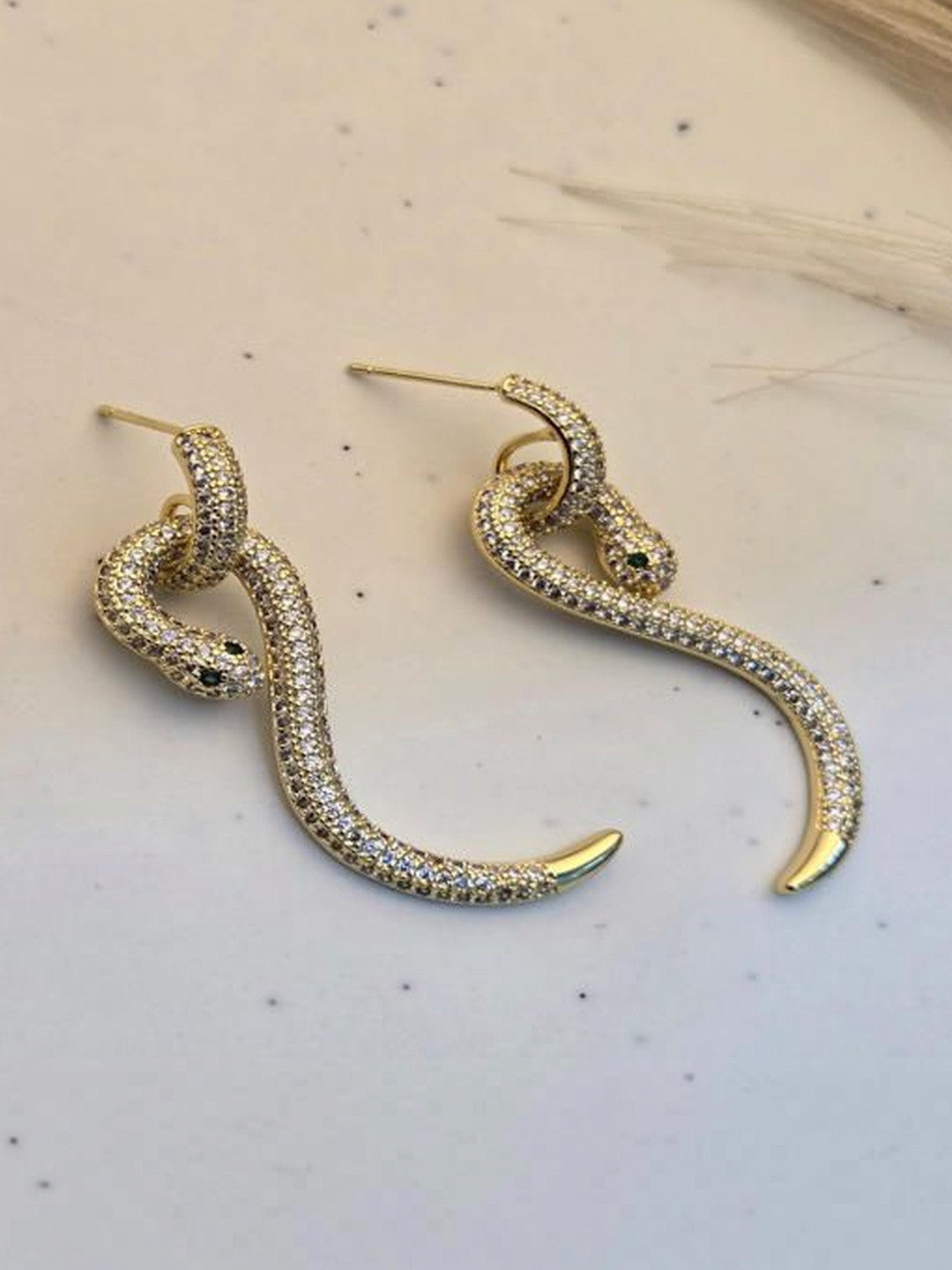 Pair of gold-plated pavé CZ snake drop earrings with green crystal eyes laid flat on a white surface