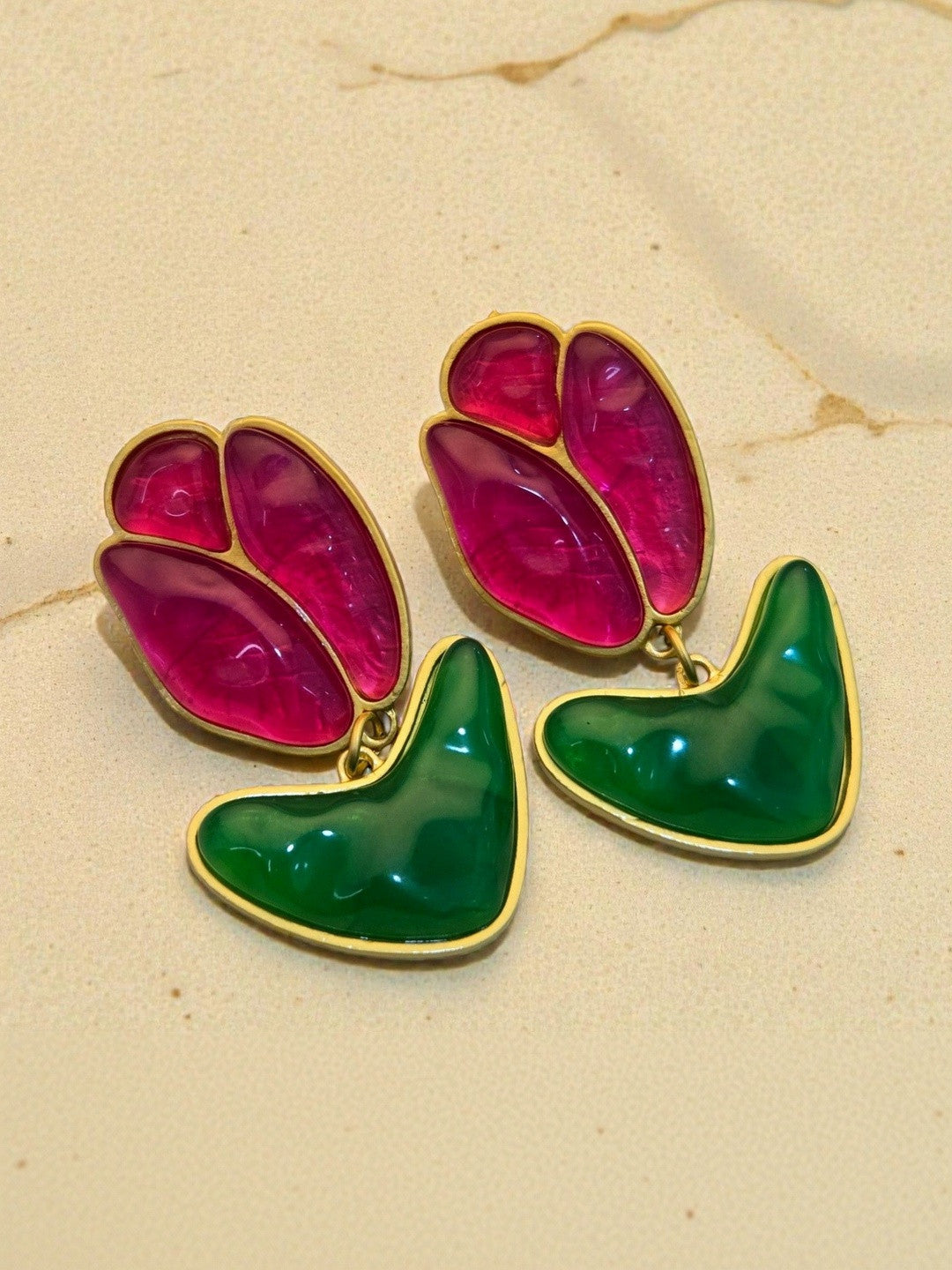 Close-up of translucent fuchsia petal and glossy green leaf gold-toned earrings on a warm beige surface