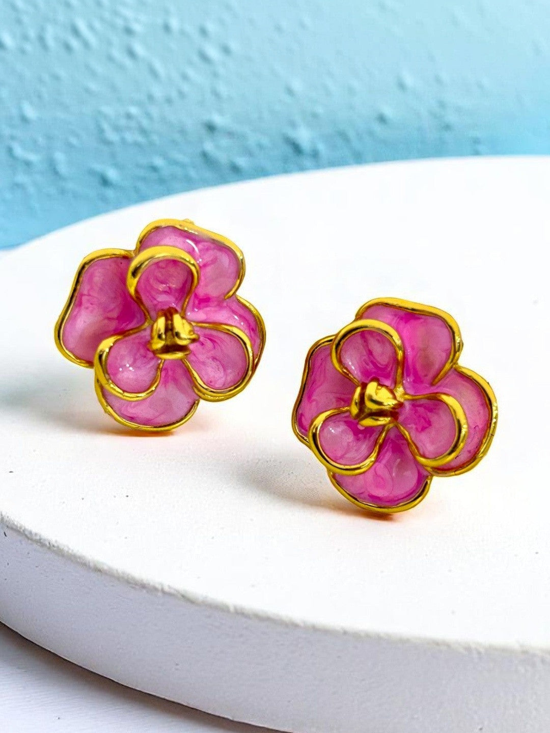 Two pink enamel flower stud earrings with gold-tone frames displayed on a white textured stand against a blue wall
