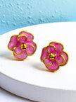 Two pink enamel flower stud earrings with gold-tone frames displayed on a white textured stand against a blue wall