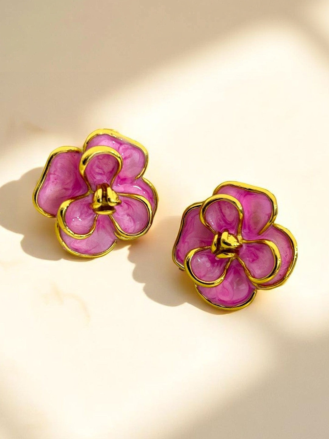 Close-up of gold-plated marbled pink enamel flower stud earrings on a warm cream surface with soft shadows