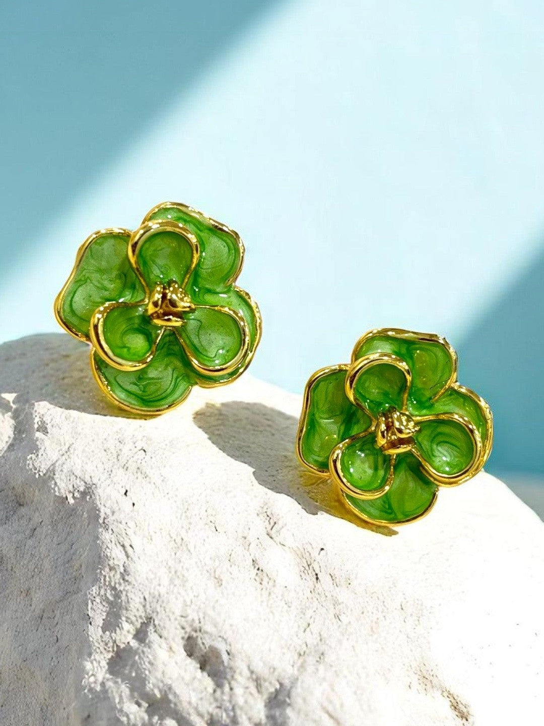 Pair of green marbled enamel floral stud earrings with gold-tone borders resting on white textured stone