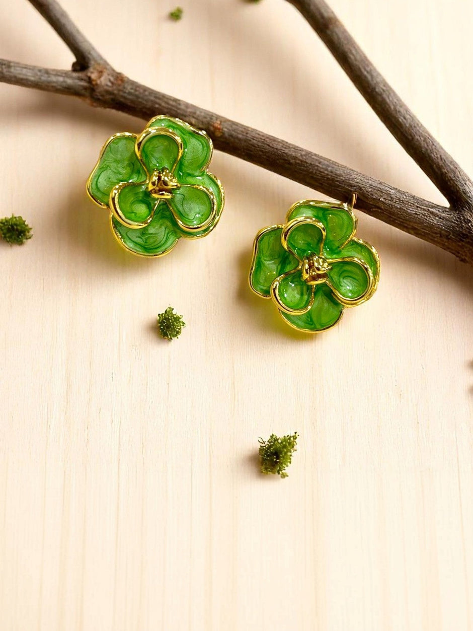 Two green enamel flower stud earrings placed beside a wooden branch with scattered moss on light wood surface