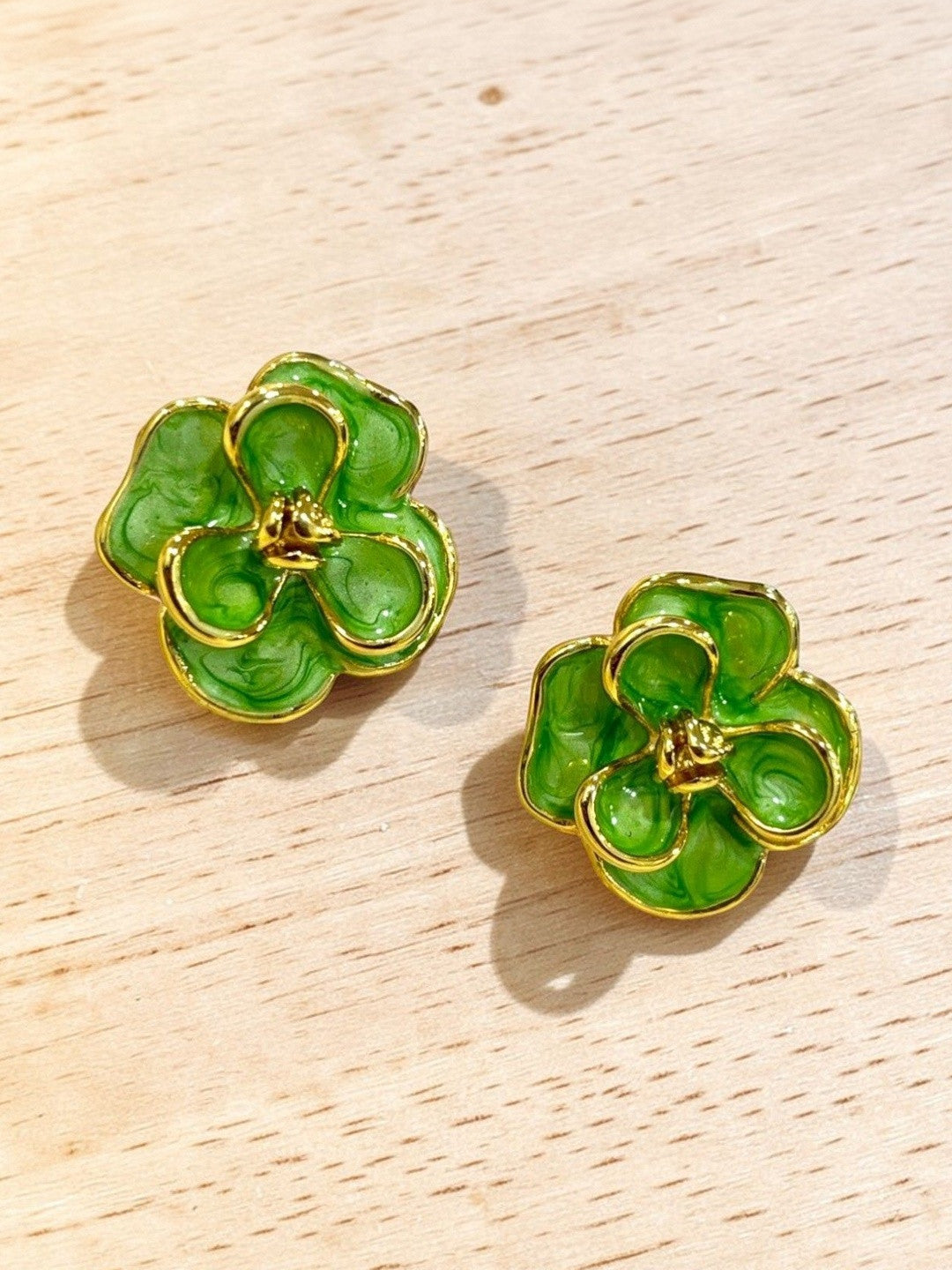 Front-facing view of green enamel gold-tone floral stud earrings lying flat on a light wooden surface