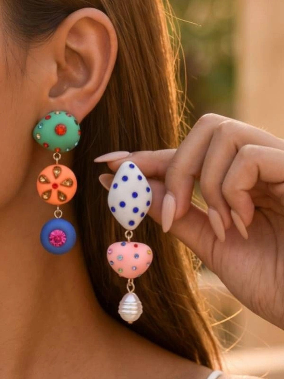 Close-up of colourful mismatched drop earrings worn on model's ear showing teal, orange, and blue stacked charms with rhinestones