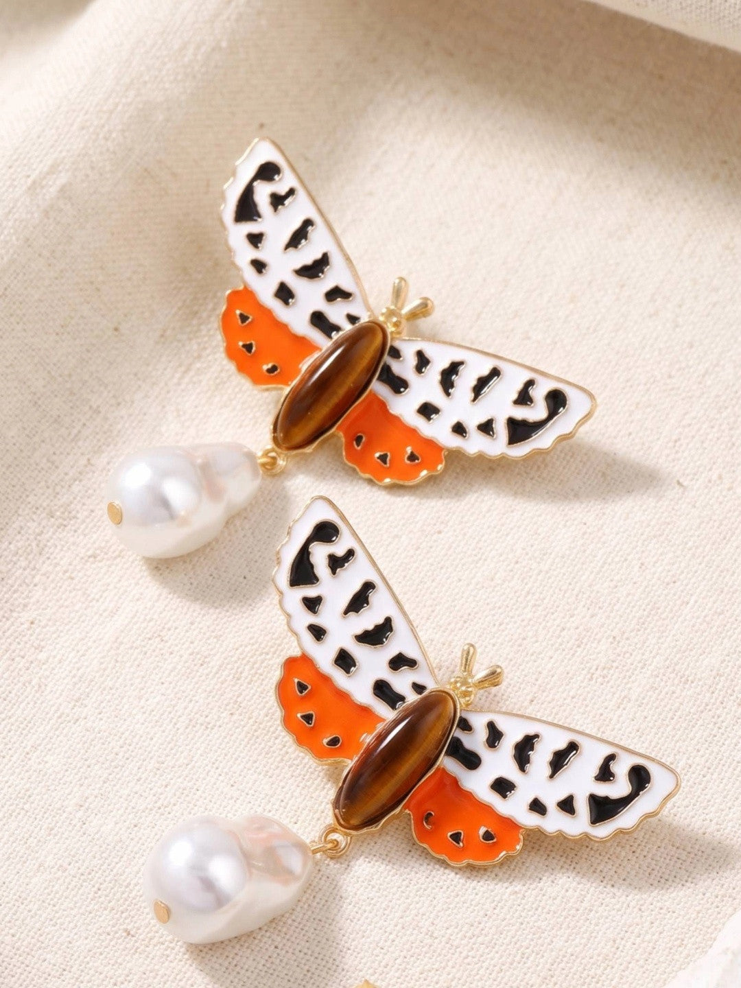 Two moth drop earrings laid flat on cream linen showing orange enamel lower wings amber oval stone and pearl drop