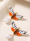 Two moth drop earrings laid flat on cream linen showing orange enamel lower wings amber oval stone and pearl drop