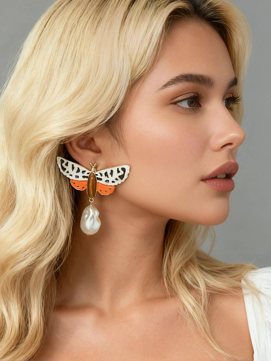 Blonde model wearing single large moth enamel drop earring with baroque pearl against grey background