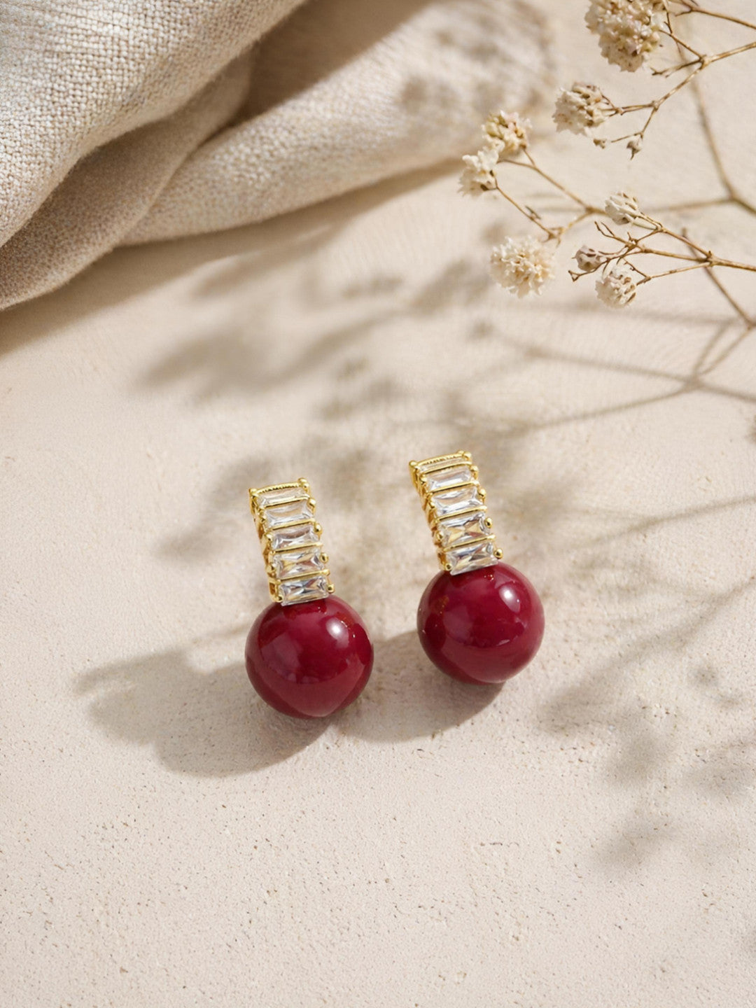 Pair of gold-plated baguette crystal bar and deep red ball drop stud earrings laid flat on a sandy beige surface with dried florals