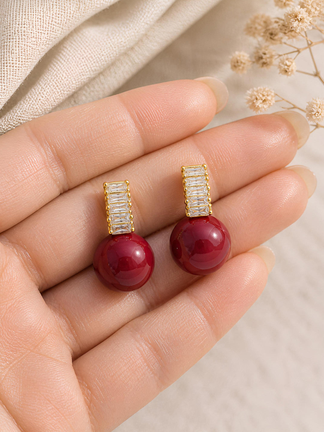 Close-up of two red ball drop stud earrings with baguette crystal tops held in an open palm to show scale