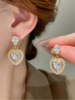 Gold-plated heart drop earring worn on ear beside a matching earring held between fingers