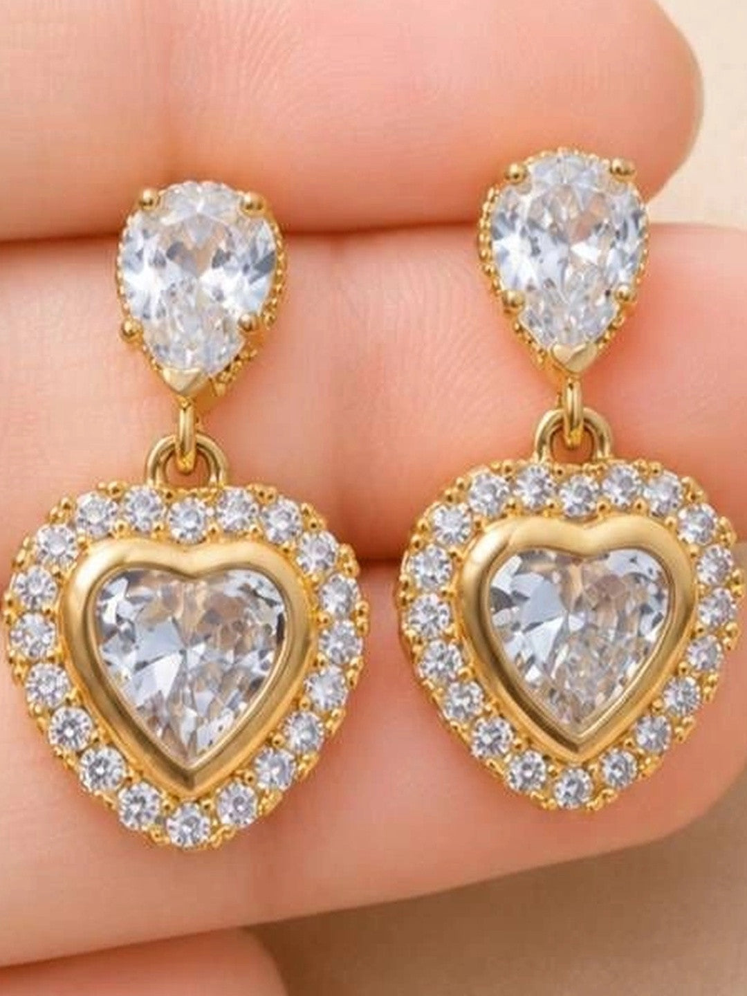 Close-up of gold heart drop earrings held in palm showing pavé CZ halo and teardrop stud