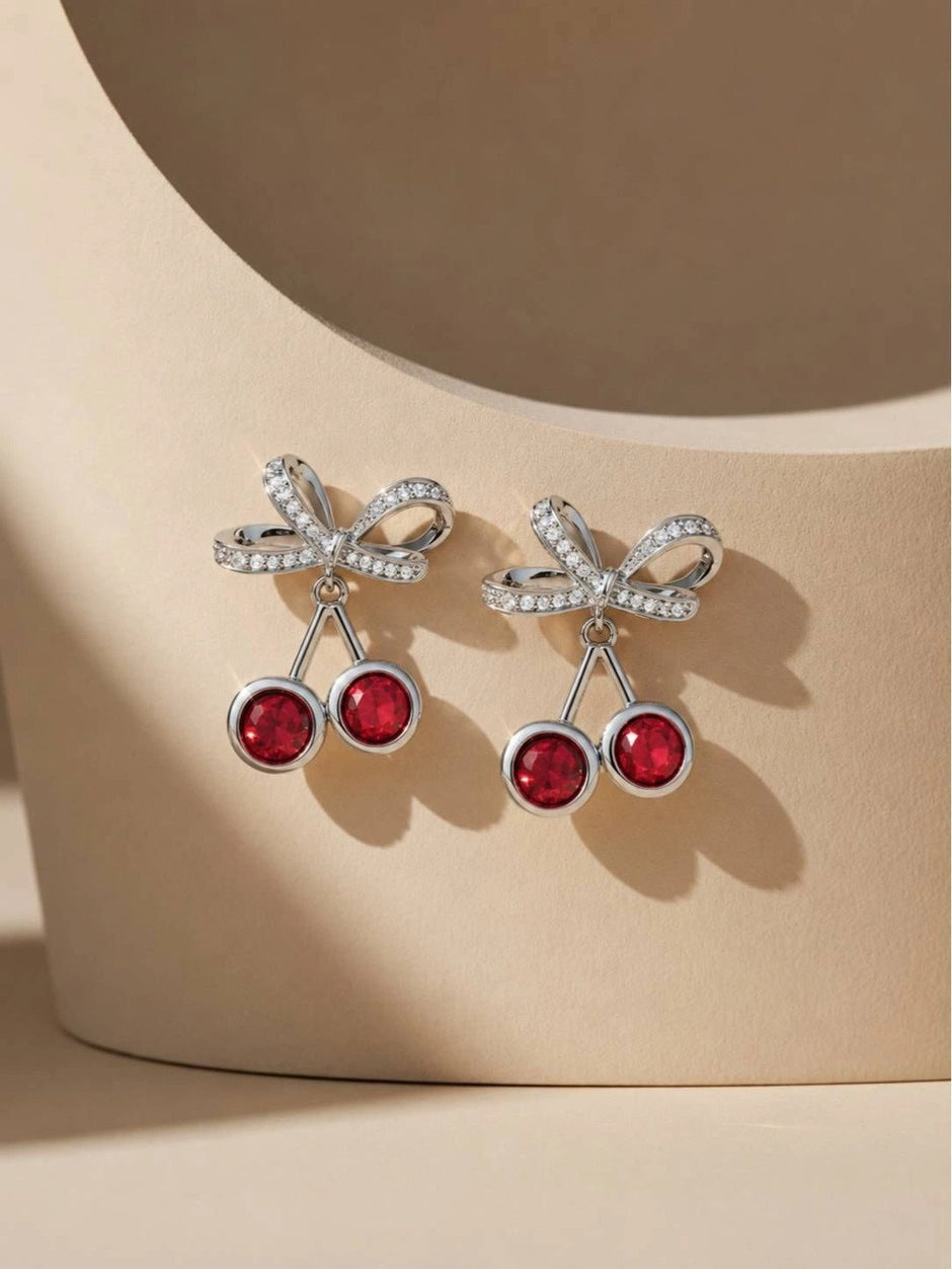 Pair of silver-tone crystal cherry bow drop earrings resting on a curved beige display prop