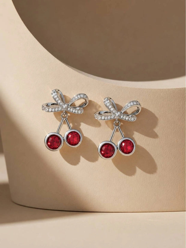 Pair of silver-tone crystal cherry bow drop earrings resting on a curved beige display prop