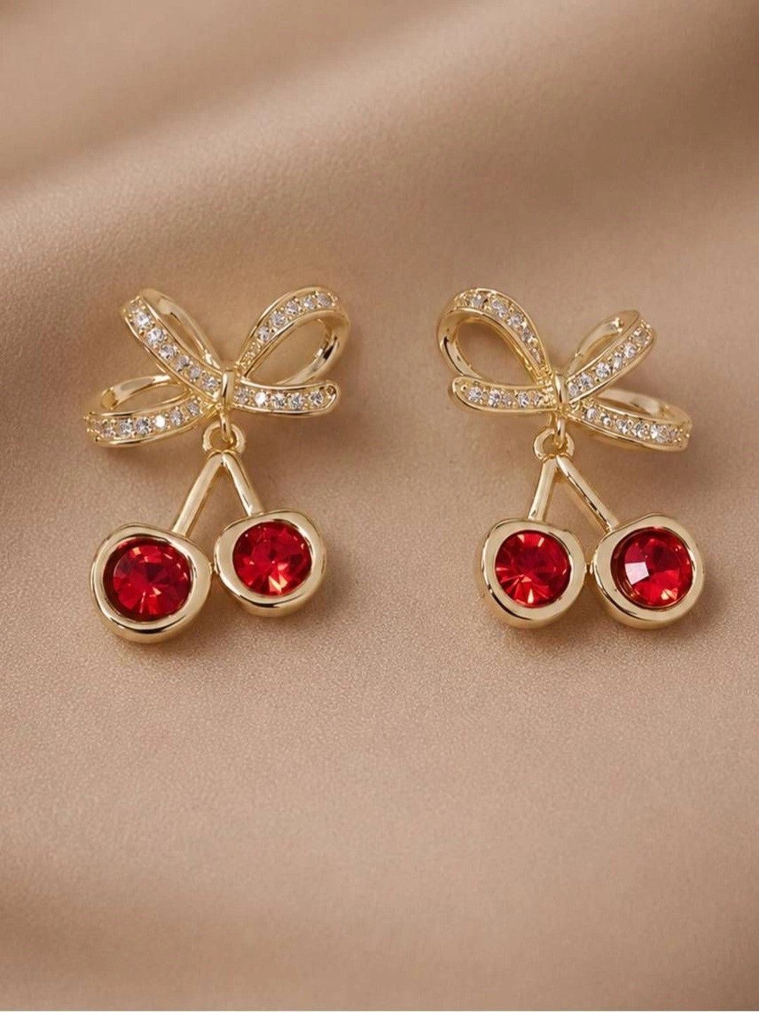 Pair of gold-plated cherry earrings with red faceted crystals and pavé bow lying flat on satin