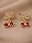 Pair of gold-plated cherry earrings with red faceted crystals and pavé bow lying flat on satin
