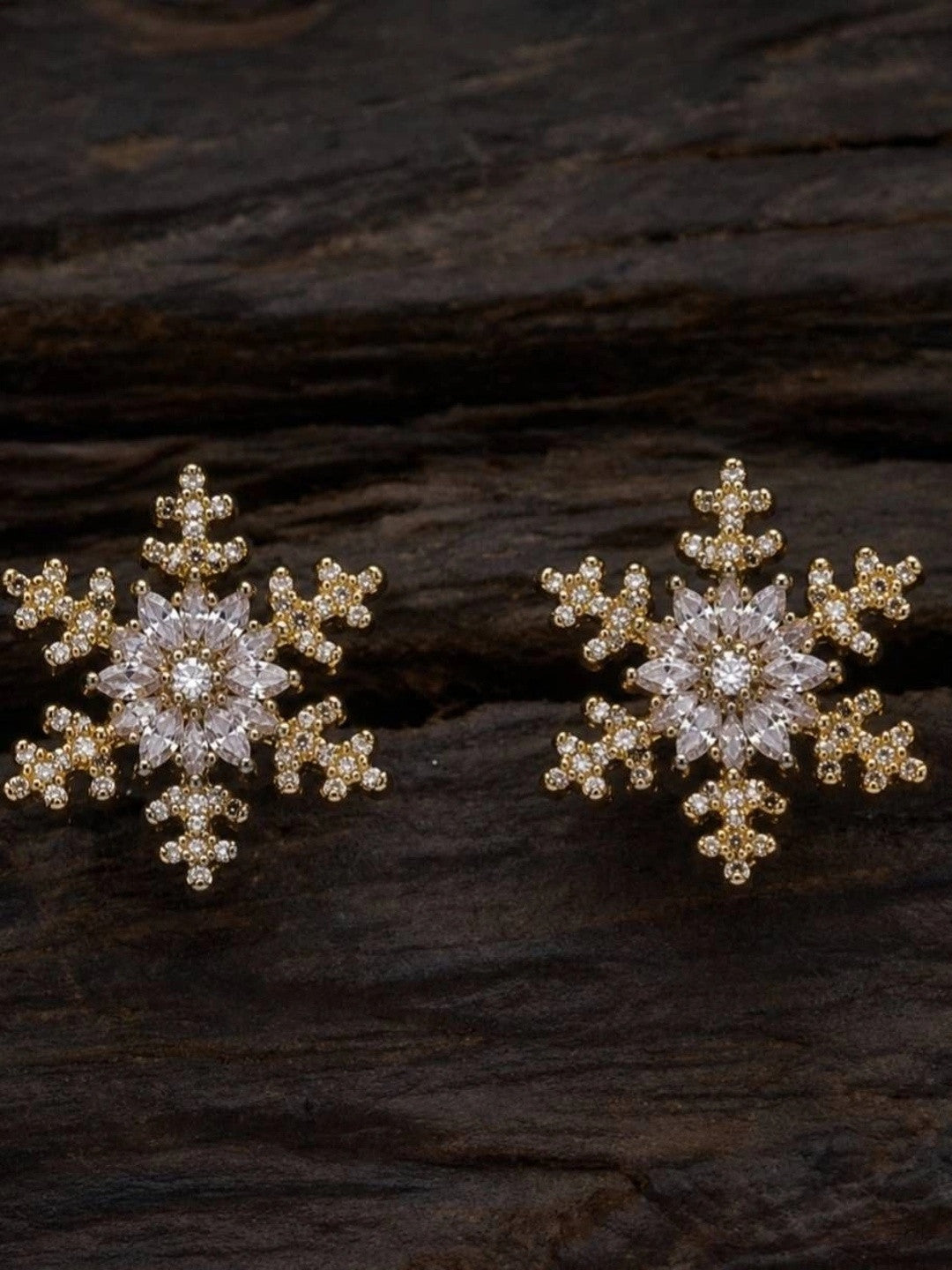 Two gold snowflake CZ stud earrings displayed on dark textured wood surface