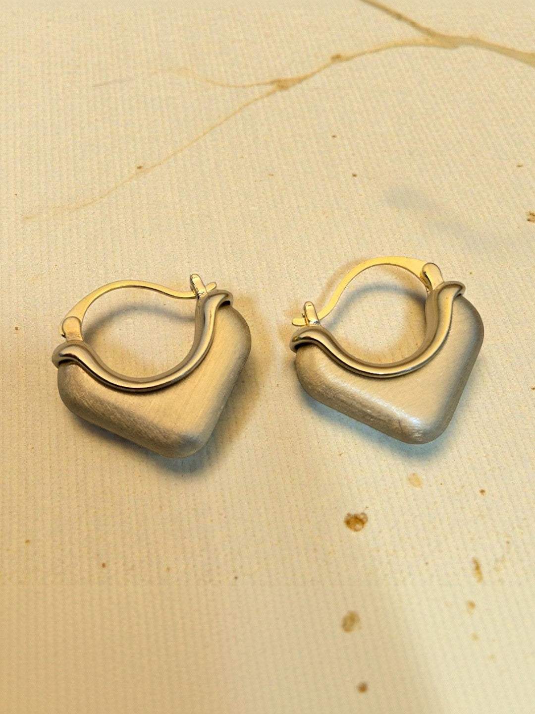 Two gold-tone square hoop earrings with click-top clasp laid flat on beige textured background