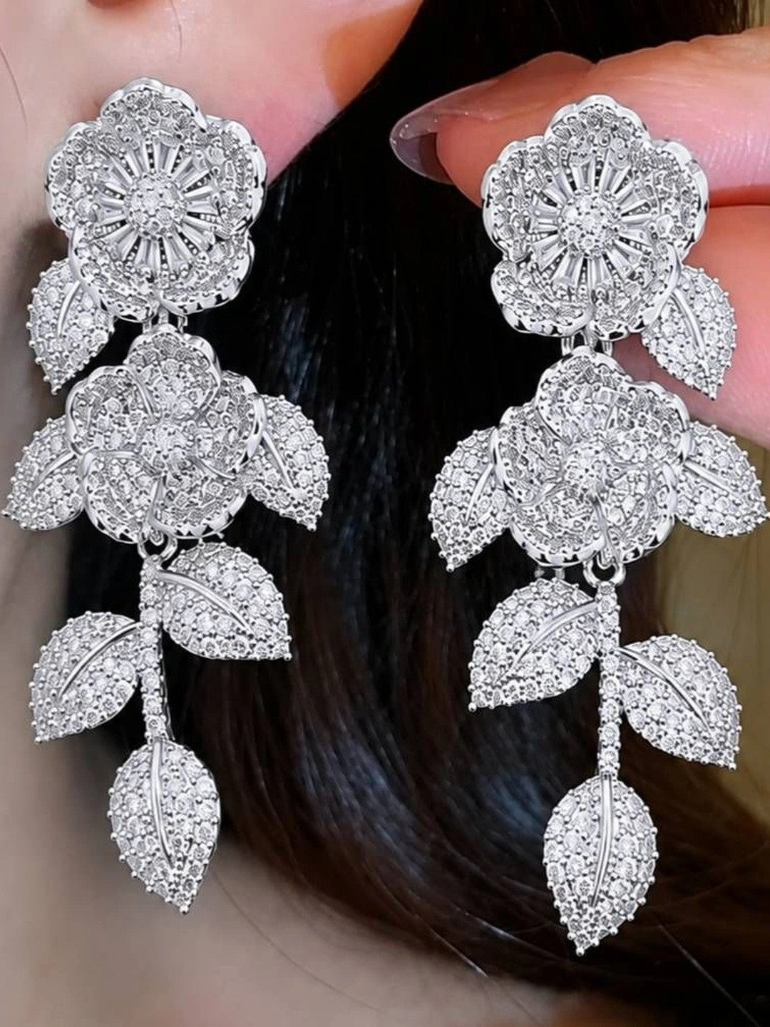 Silver CZ floral drop earrings held near a model's ear showing wearable drape and scale