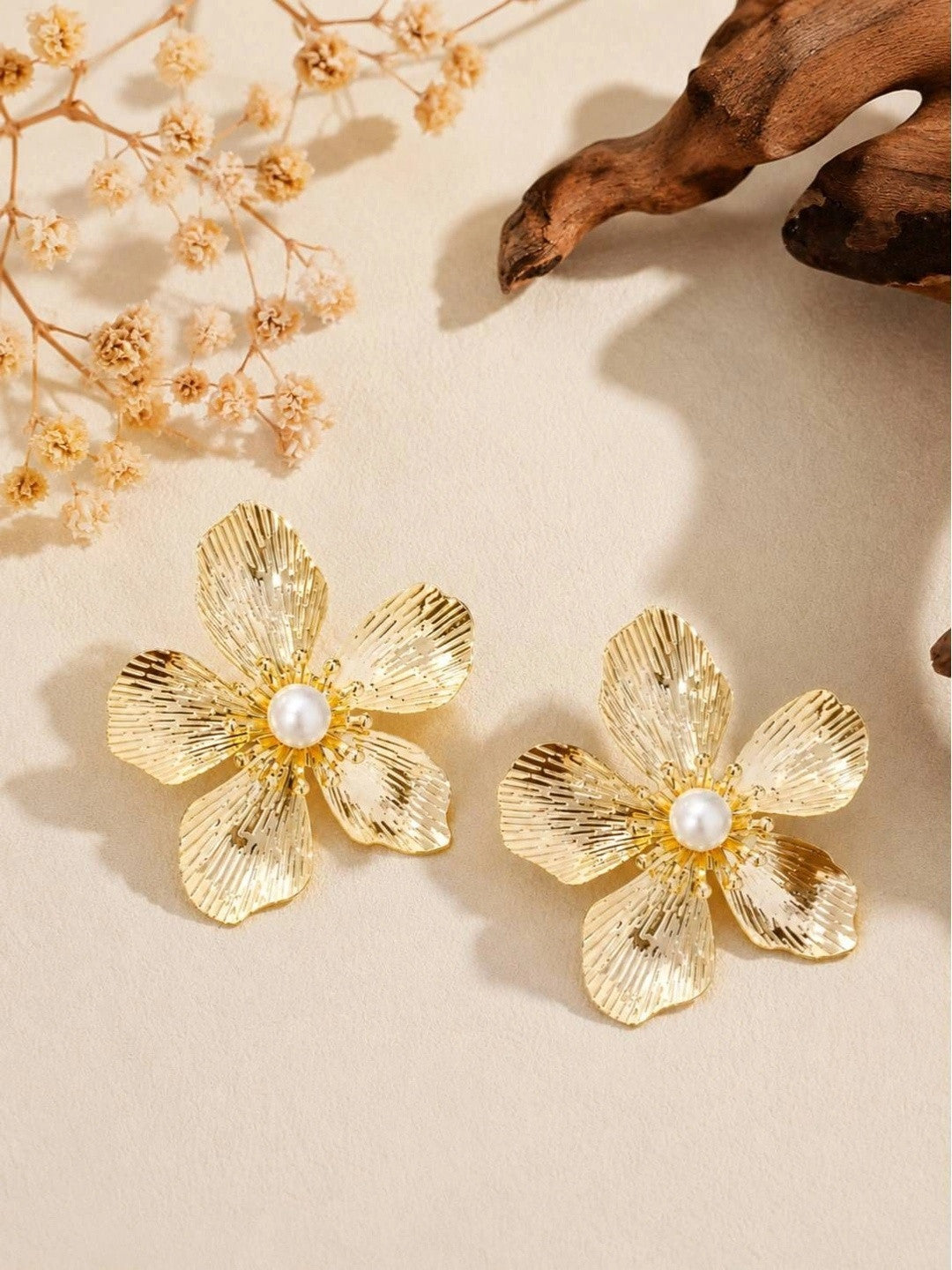 Pair of gold-toned five-petal flower stud earrings with faux pearl centres on a beige surface with dried flowers