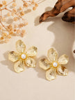 Pair of gold-toned five-petal flower stud earrings with faux pearl centres on a beige surface with dried flowers