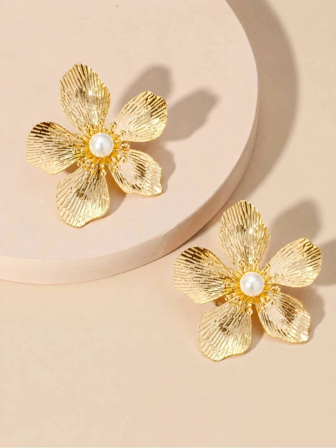 Two gold ribbed floral stud earrings displayed on a round white platform against a warm beige background