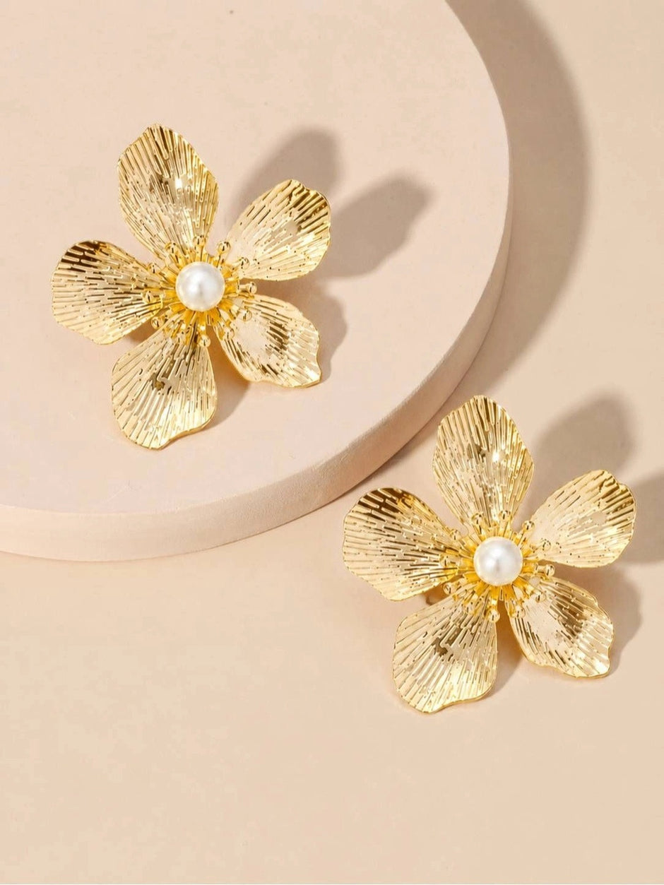 Two gold ribbed floral stud earrings displayed on a round white platform against a warm beige background