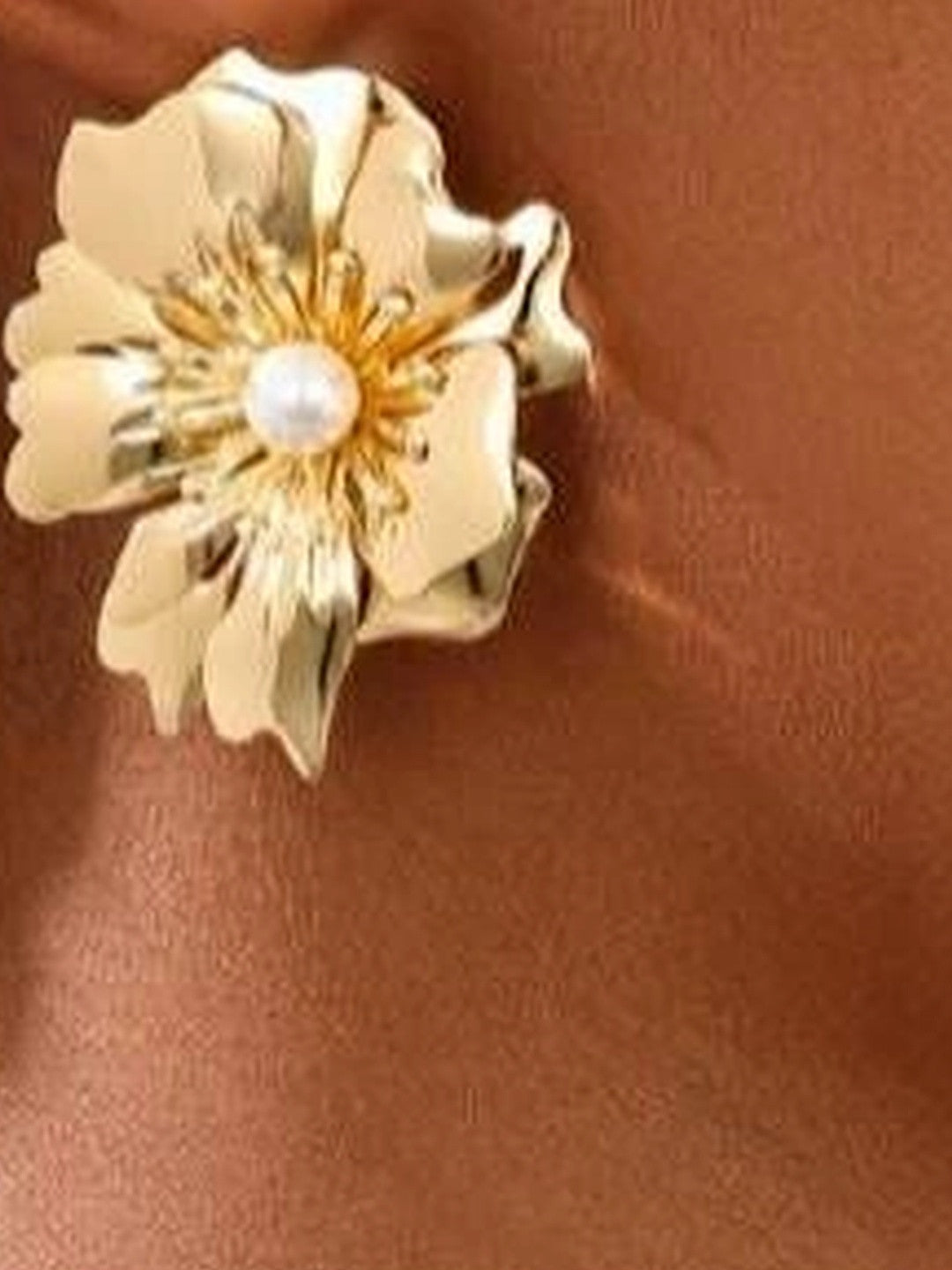 Gold floral stud earring with pearl centre worn on a model's ear against a warm brown background