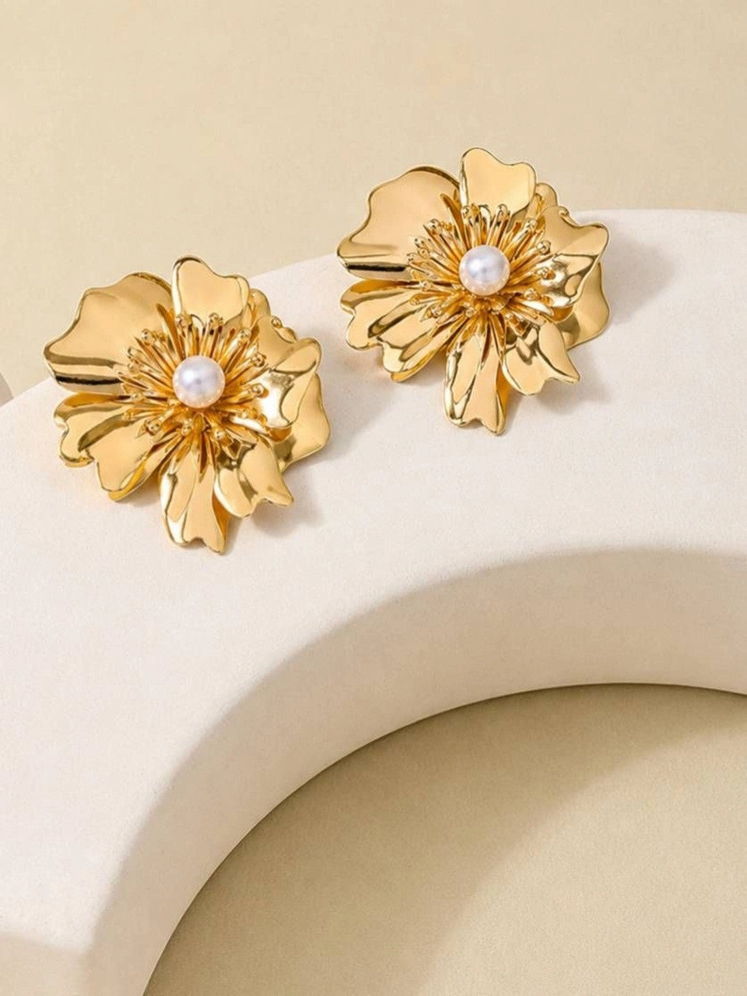 Pair of gold-plated layered petal stud earrings with pearl centres displayed on a curved white prop