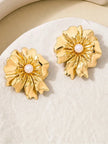 Two gold floral stud earrings with ruffled petals and pearl centres resting on a textured stone surface