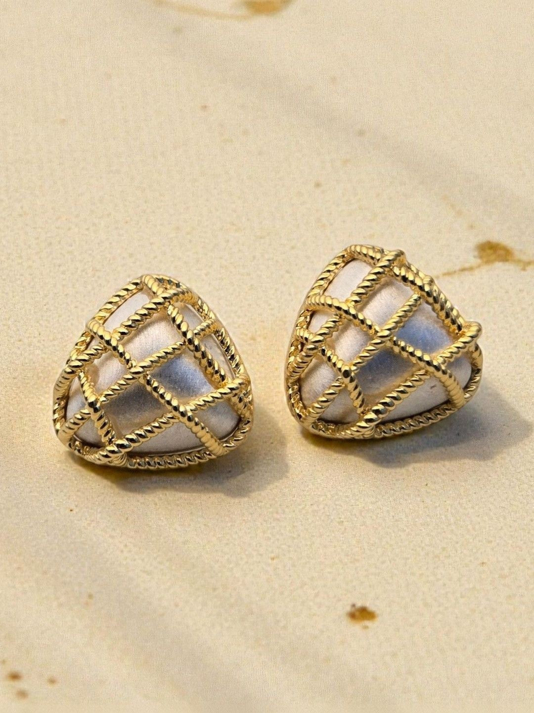 Close-up of two gold rope-grid heart studs with white pearl inlay on a textured beige surface