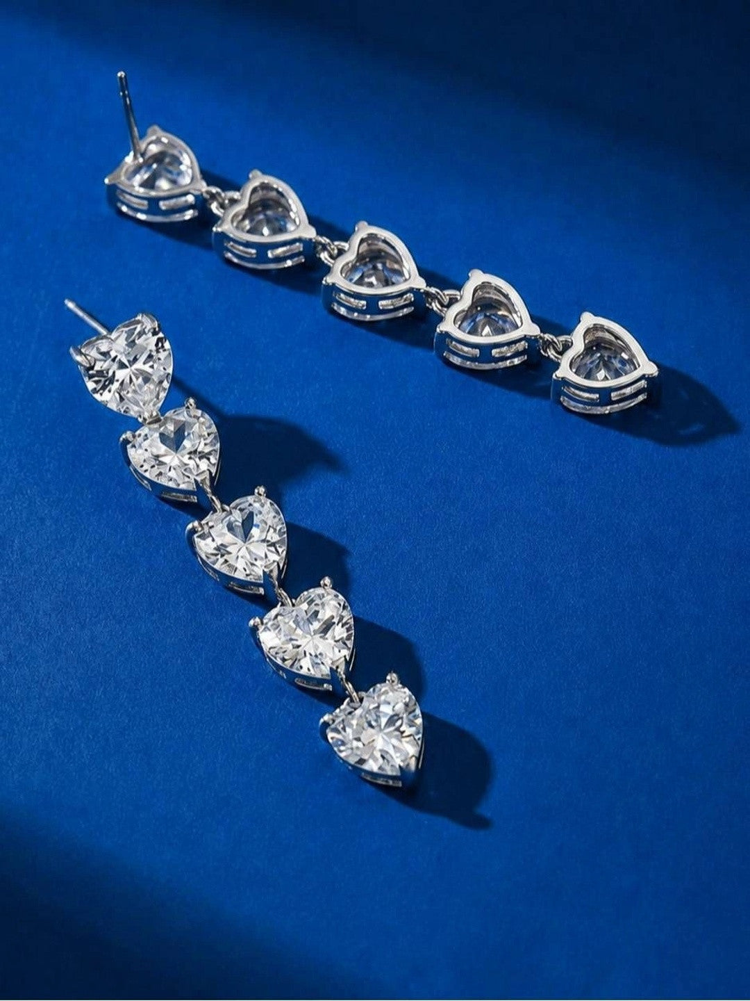 Two silver-finish heart drop earrings laid flat on a royal blue surface, showing the back and front side by side