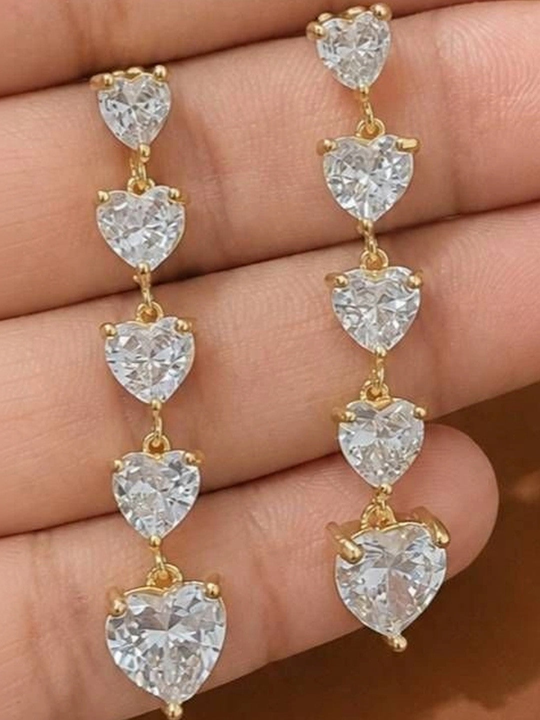 Close-up of heart CZ drop earrings held in a palm, highlighting five linked cubic zirconia hearts and gold connecting links