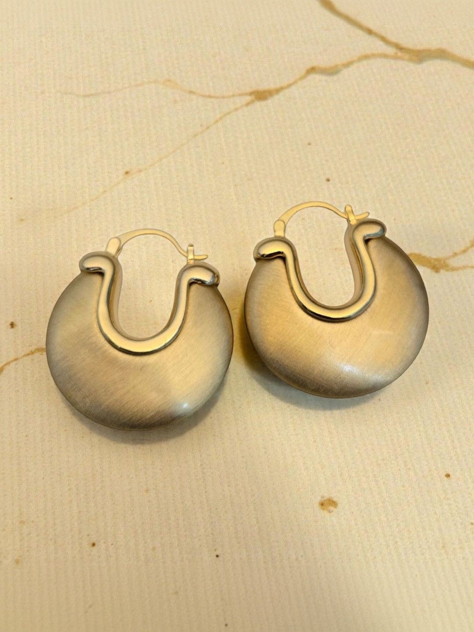 Close-up of two matte gold disc hoop earrings on a beige paper surface showing brushed texture