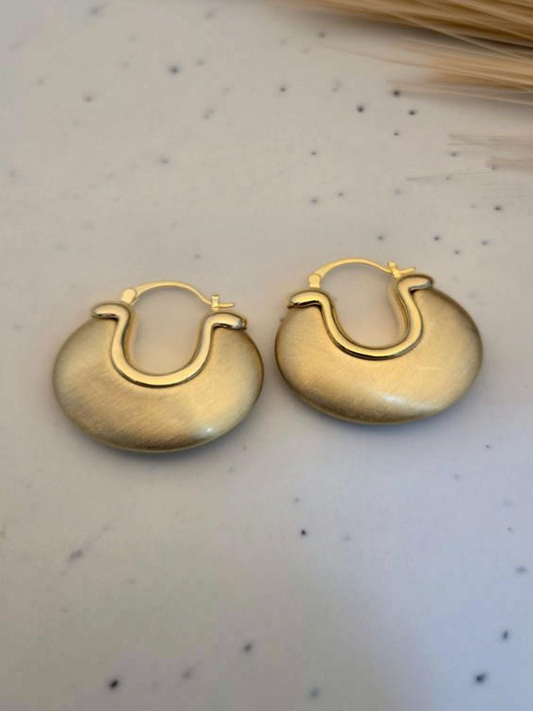 Pair of matte gold chunky disc hoops placed on a white speckled marble surface near dried grass